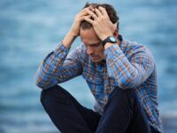 A man in a plaid shirt sits by the water looking distressed, symbolizing stress.